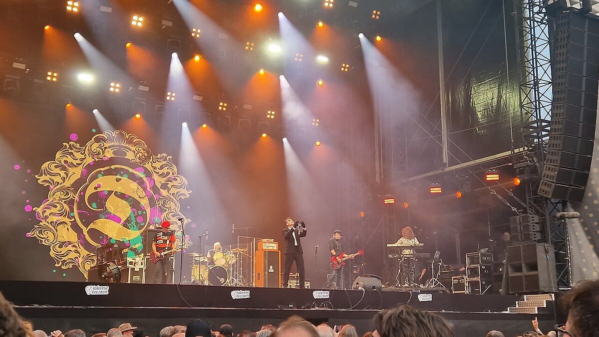 The Damned performing on a festival stage under orange and white spotlights, with the band's gold logo on a backdrop