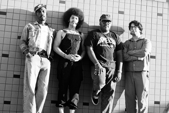 Four members of a band standing against a white tiled wall in a black and white photo