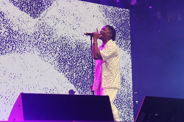 Lucky Daye singing into a microphone on a stage lit in purple, in front of a large video screen