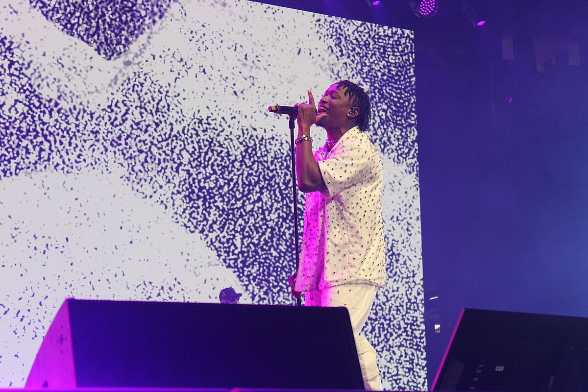 Lucky Daye singing into a microphone on a stage lit in purple, in front of a large video screen