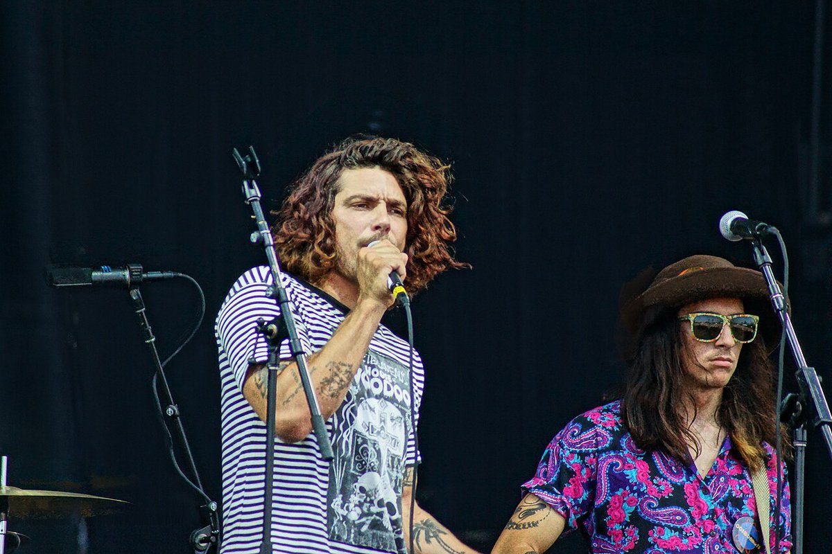 Two members of a band on stage at microphones, one singing in a striped shirt, the other in a hat and sunglasses