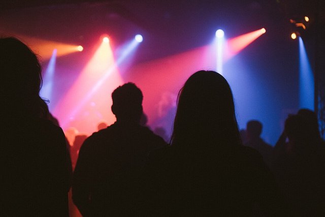 Silhouetted audience members facing a stage lit by pink, orange and blue spotlights