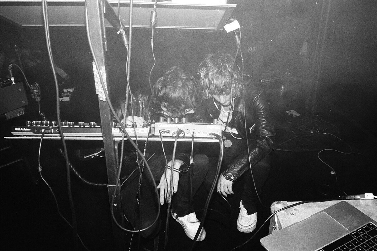 Two band members hunched over a synthesizer and drum machine on a dark stage, tangled in cables