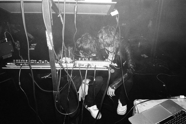 Two band members hunched over a synthesizer and drum machine on a dark stage, tangled in cables