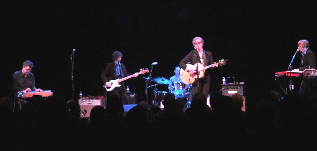 The Jayhawks on a dark stage with a singer on acoustic guitar, a bassist, a drummer and two keyboard players
