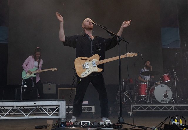 Singer-guitarist with arms raised at the microphone, with a guitarist and drummer on stage behind him