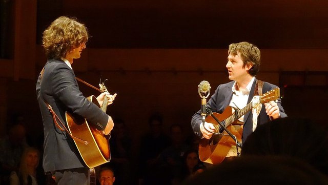 Two musicians facing each other on stage, each playing an acoustic guitar beside a vintage-style microphone