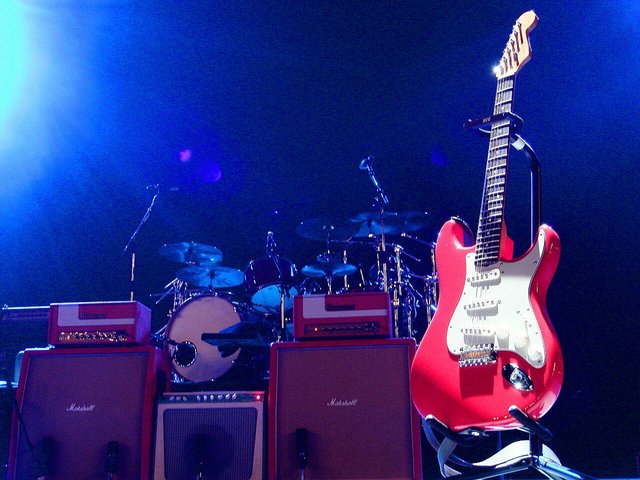 A red electric guitar standing in front of amplifiers and a drum kit under blue stage light