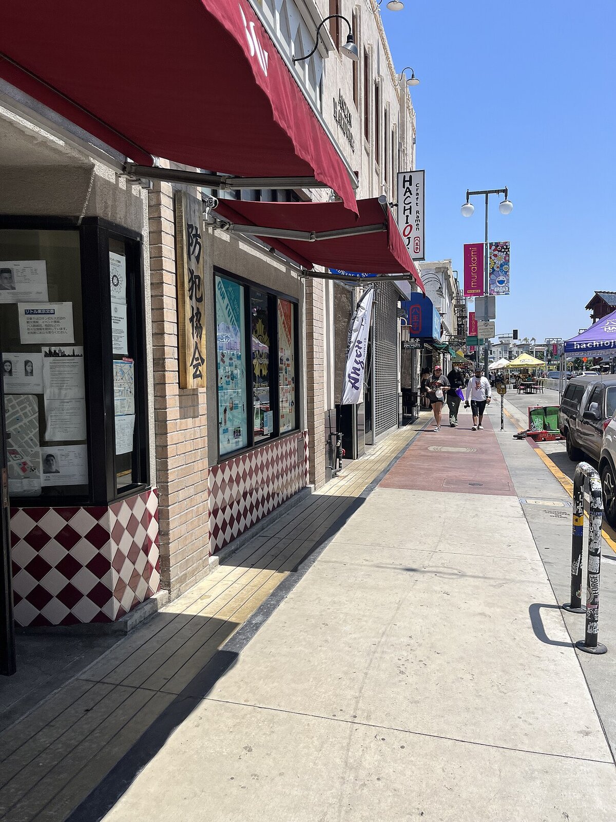 Sunlit sidewalk along 1st Street in Little Tokyo with red awnings and ramen shop signs, a few pedestrians in the distance
