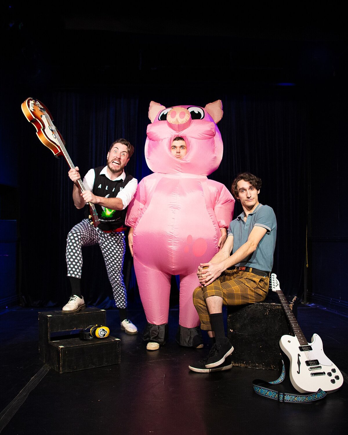 Three members of The Toxhards on a dark stage, one swinging a bass, one seated with a guitar, one in an inflatable pink pig costume