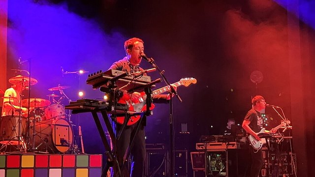 The Wombats on stage under purple and red lights, a singer at a keyboard and guitar with a drummer behind