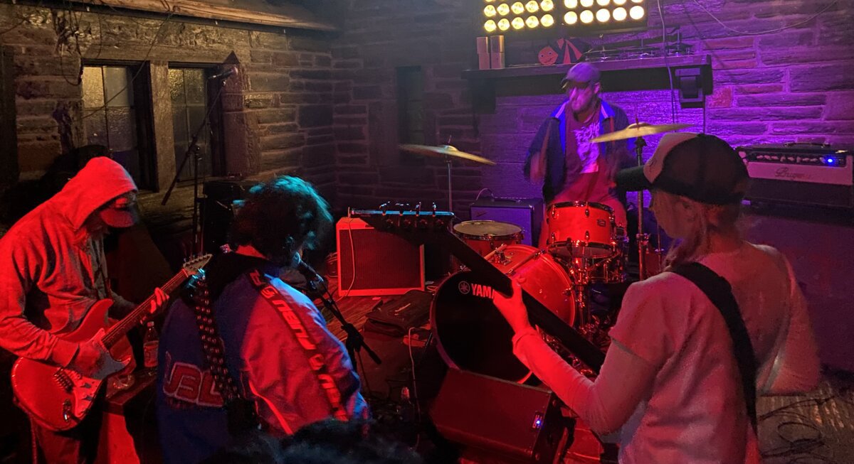 A band playing in a small brick room under red and purple stage light, with guitarists in the foreground and a drummer behind