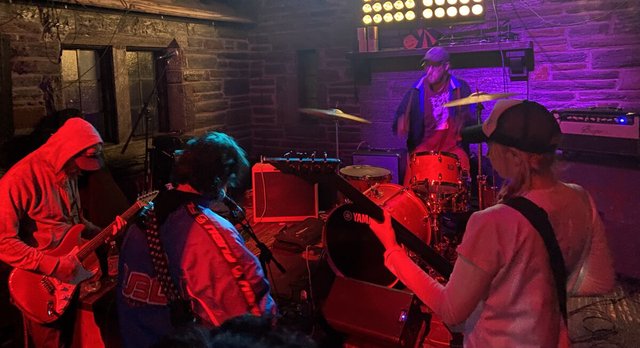 A band playing in a small brick room under red and purple stage light, with guitarists in the foreground and a drummer behind