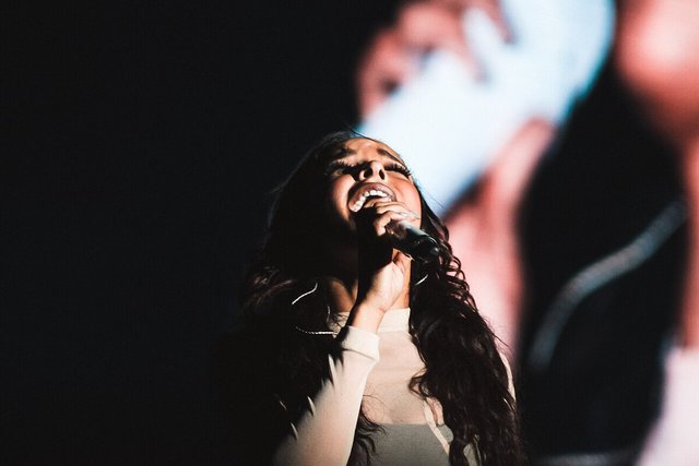 Singer Tinashe performing on stage, head tilted back, holding a microphone in front of a dark video screen