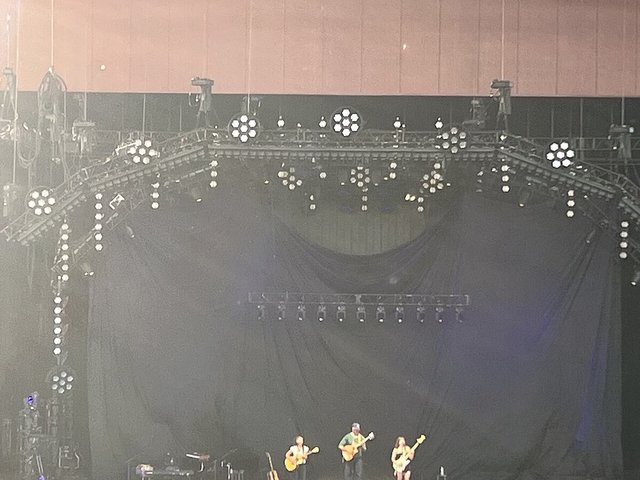 Three musicians with guitars and a bass on a large, dim stage under hanging truss lights