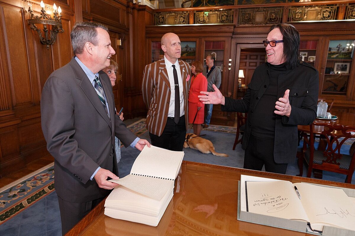 Todd Rundgren, in dark clothes and tinted glasses, gesturing mid-conversation beside a desk in a wood-paneled room
