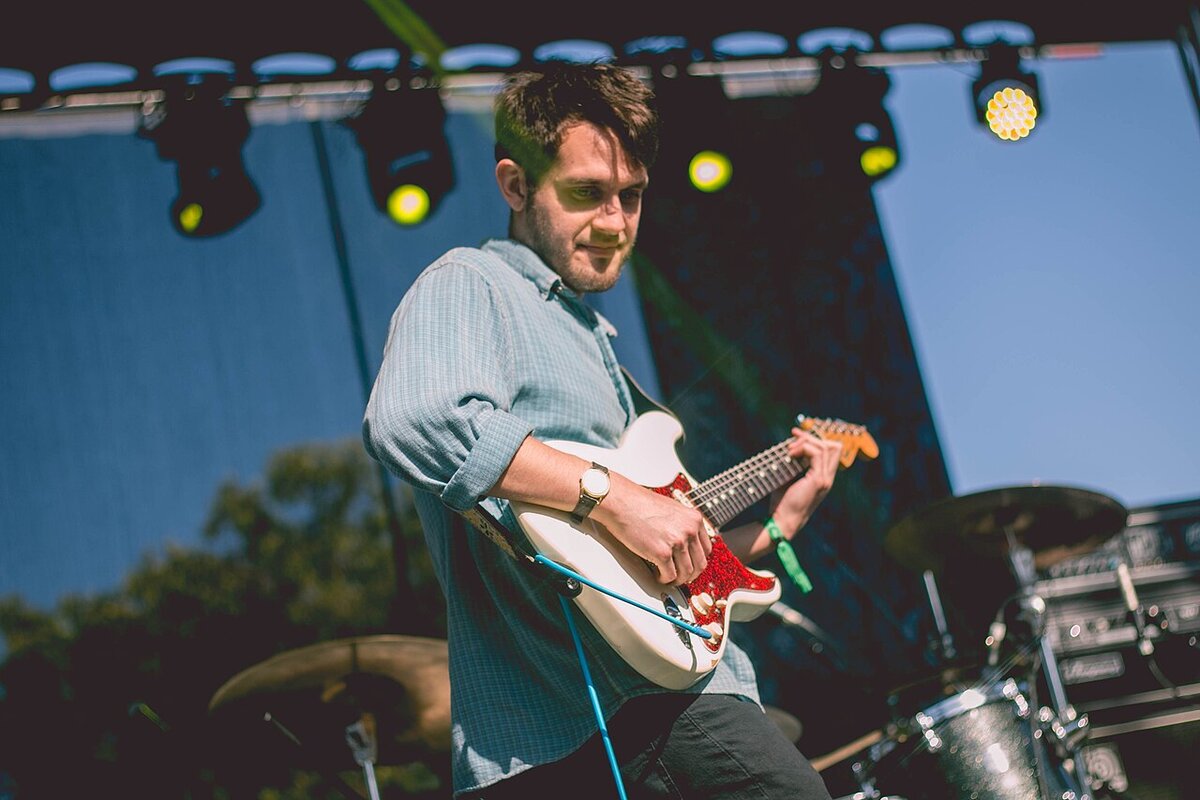 Guitarist in a button-up shirt playing a white electric guitar on an outdoor stage under stage lights