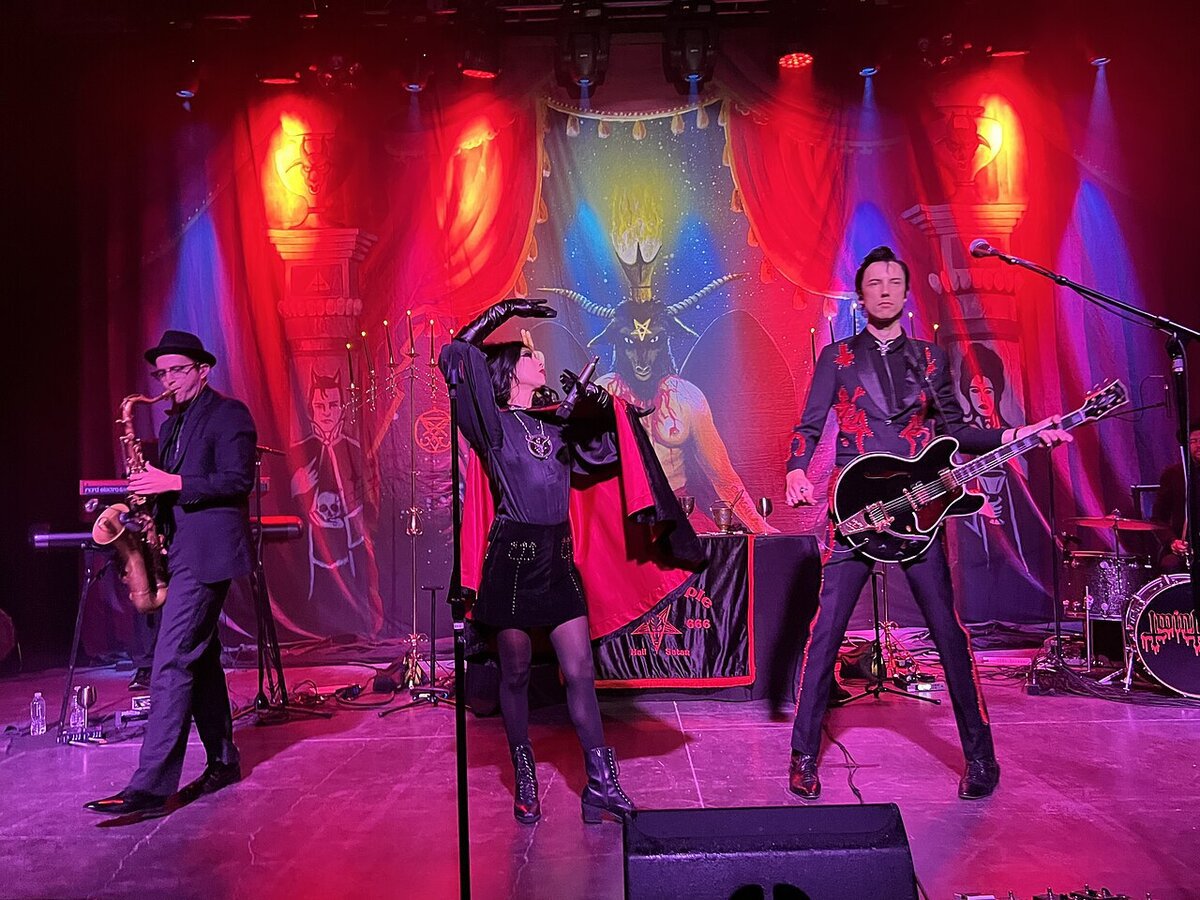 Twin Temple performing on a red-lit stage with a saxophonist, a singer in a cape and a guitarist in front of an occult-themed backdrop