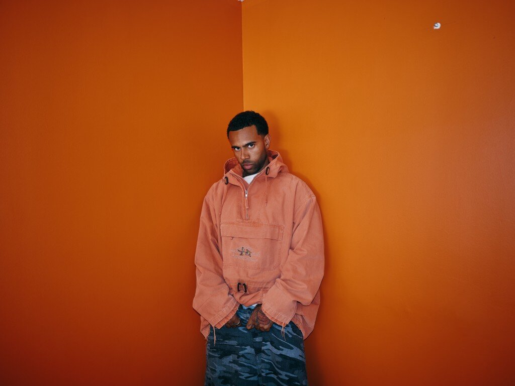 Vic Mensa in an orange anorak and camouflage pants, leaning in the corner of an orange room