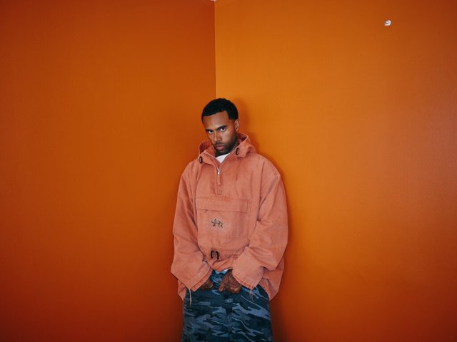 Vic Mensa in an orange anorak and camouflage pants, leaning in the corner of an orange room