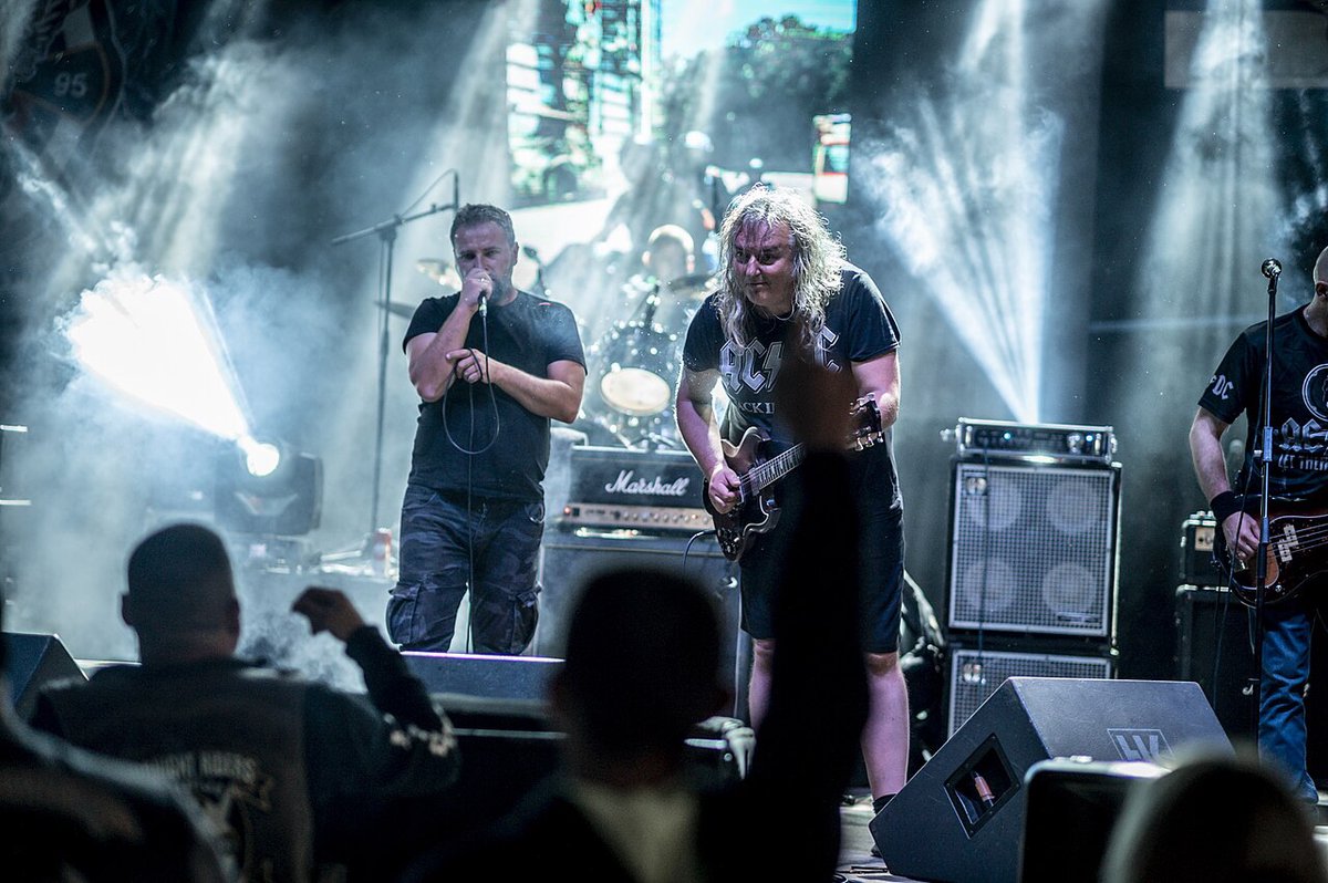 A rock band on a lit outdoor stage with a crowd in front