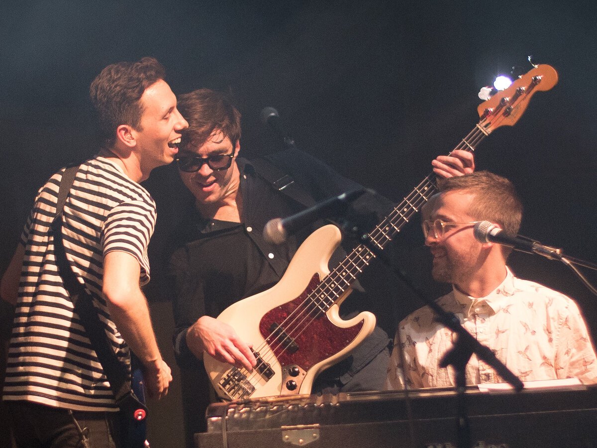 Three band members laughing together on a dark stage, one in a striped shirt, one playing a cream bass guitar, one at a keyboard