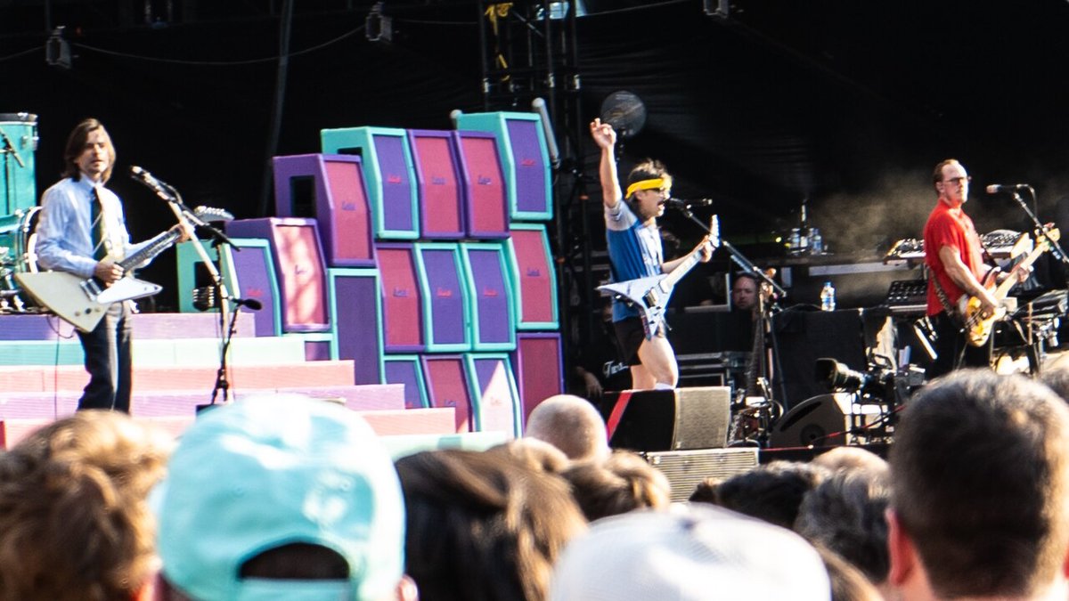 Weezer on an outdoor stage in front of stacked teal, purple and pink guitar amps, Rivers Cuomo in a yellow headband with his arm raised
