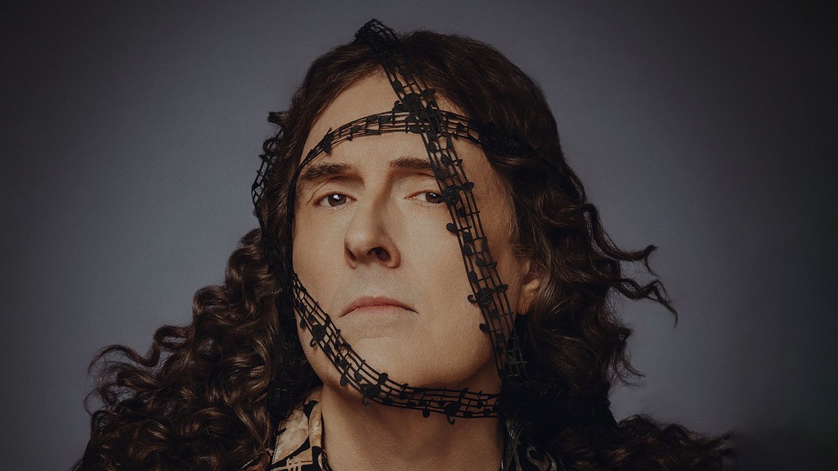 "Weird Al" Yankovic with long curly hair, a ribbon of black music notes wrapped across his face