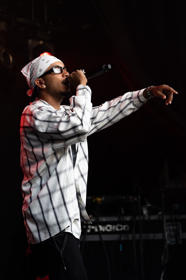 Rapper in a white bandana and checked shirt performing on stage with a microphone, arm extended