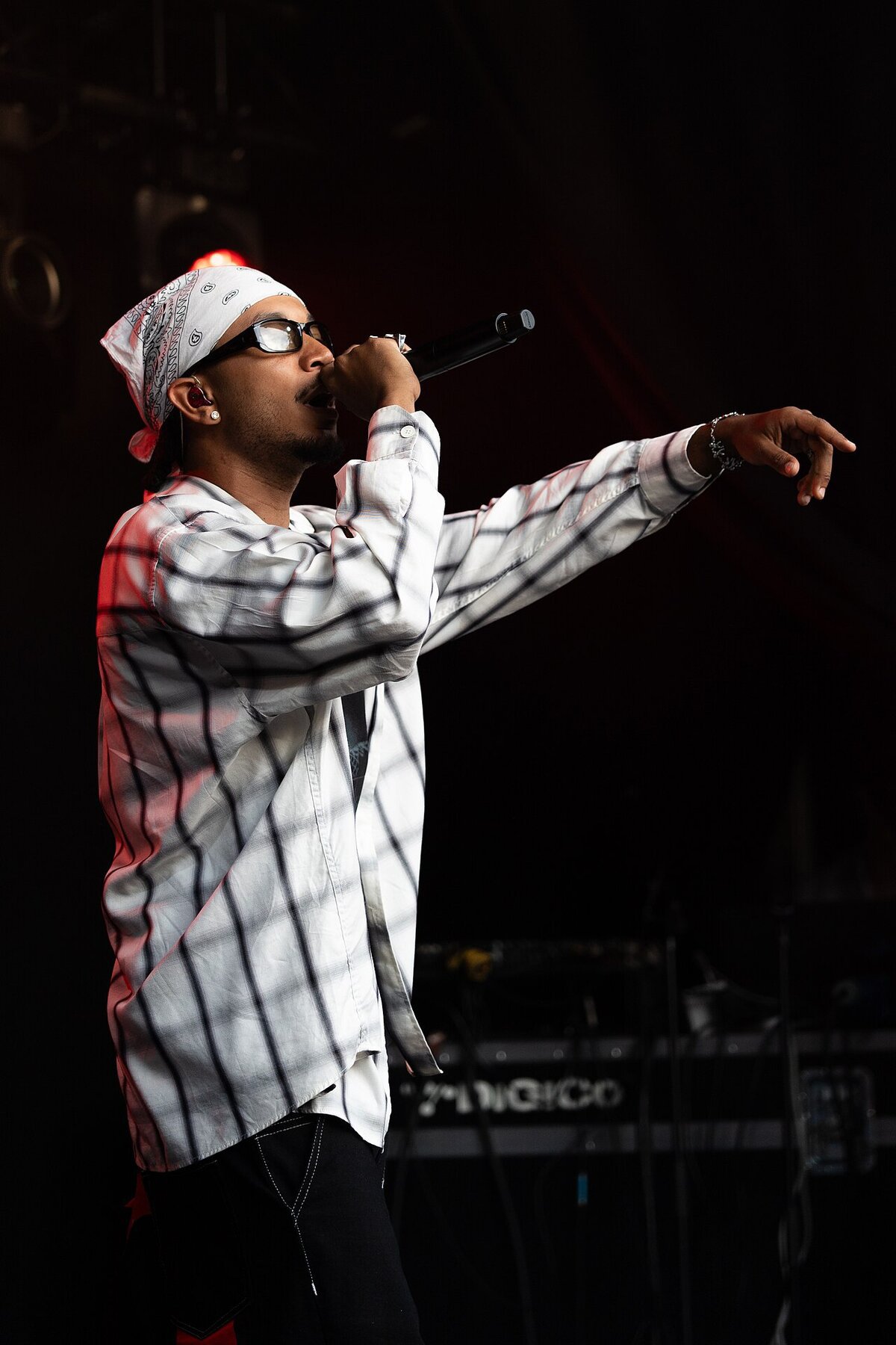 Rapper in a white bandana and checked shirt performing on stage with a microphone, arm extended