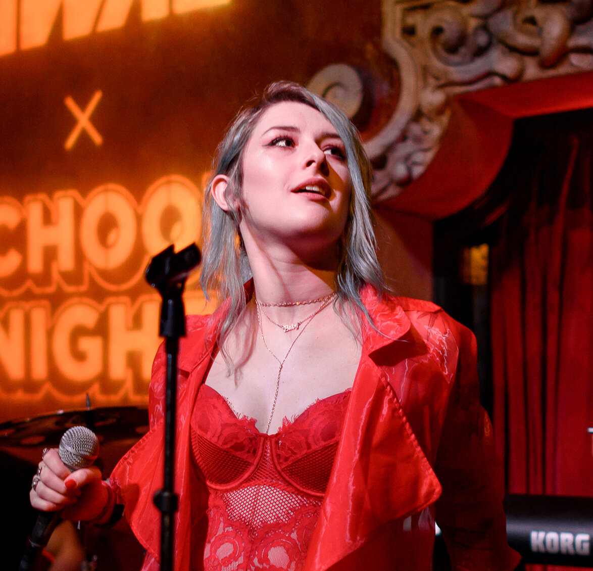 Singer with pale blue hair in a red jacket holding a microphone on a stage with warm orange lighting