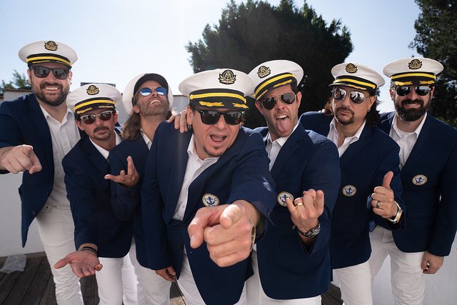 Seven members of Yachtley Crew in navy blazers and captain's hats, pointing at the camera
