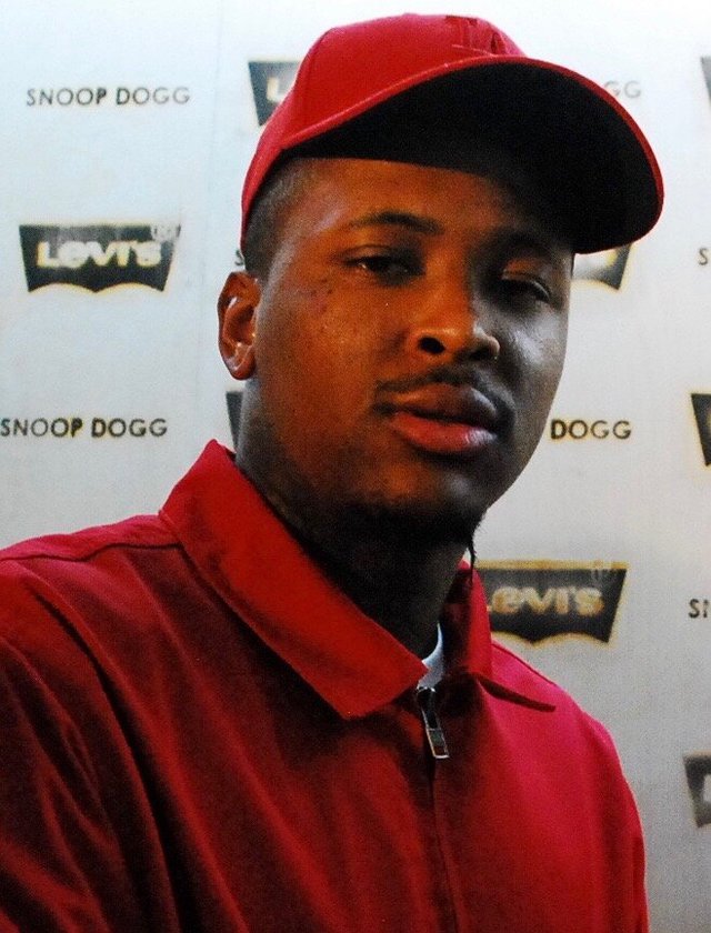 Portrait of rapper YG wearing a red cap and red jacket in front of a step-and-repeat backdrop