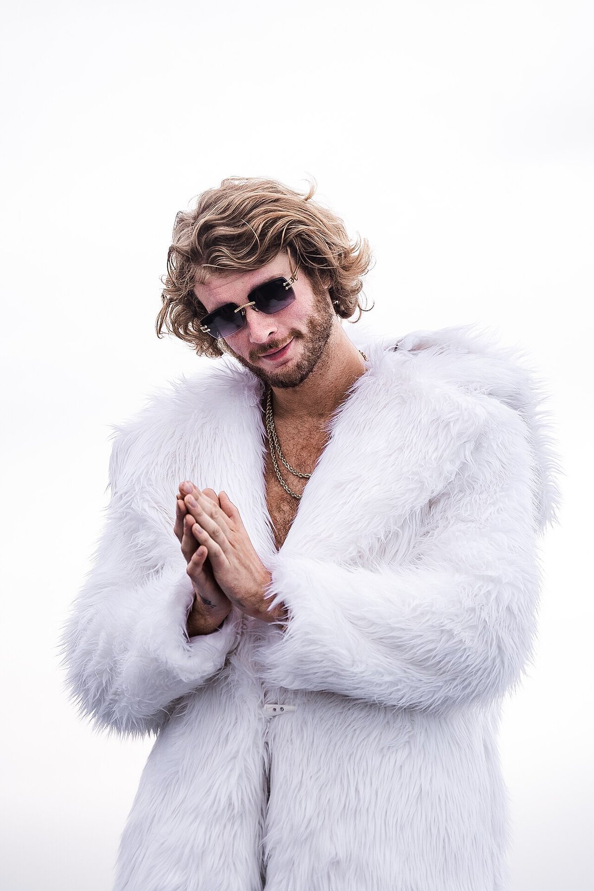 Yung Gravy in sunglasses and a white fur coat, hands pressed together, against a pale sky