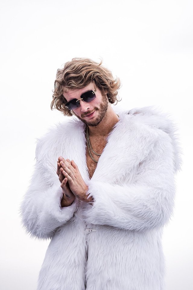 Yung Gravy in sunglasses and a white fur coat, hands pressed together, against a pale sky