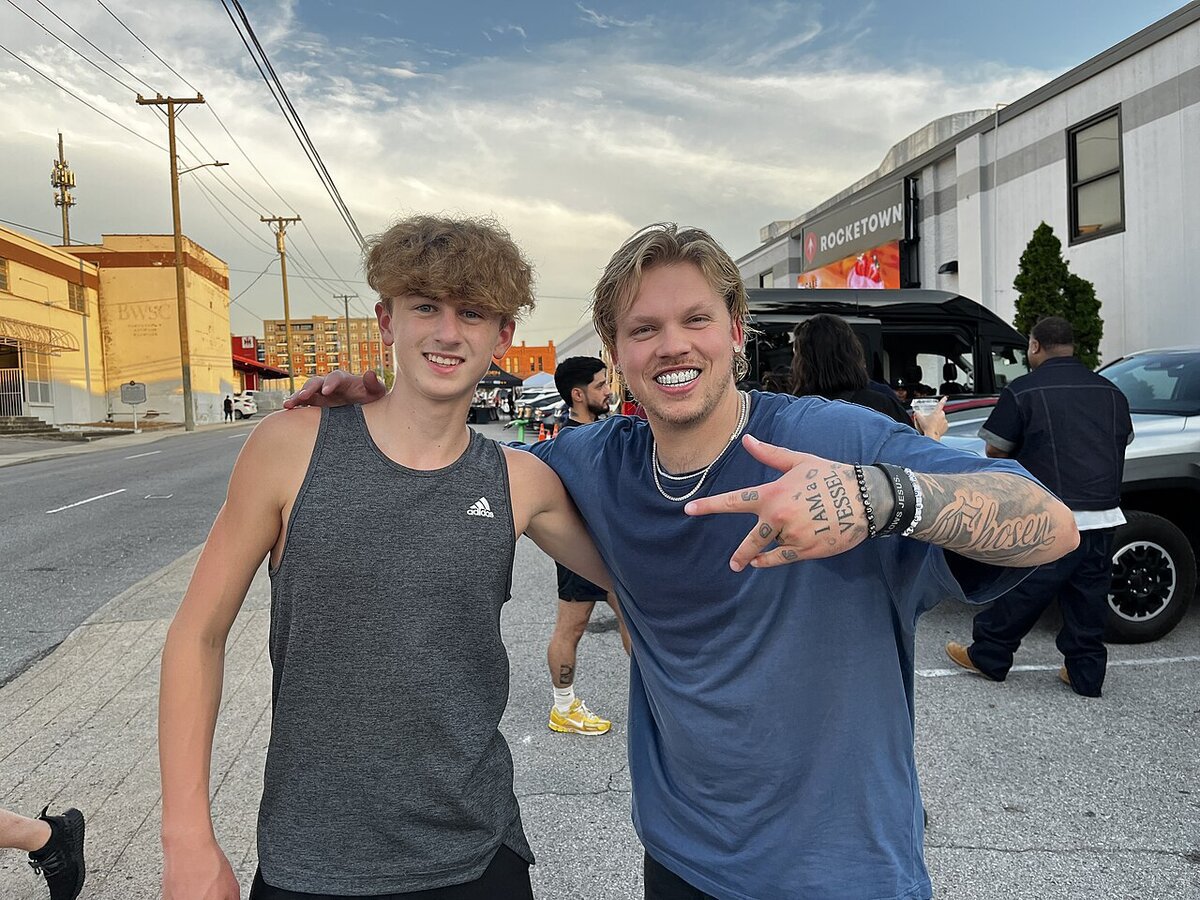 Two men posing for a photo on a city street, the one on the right wearing a blue t-shirt and pointing at the camera