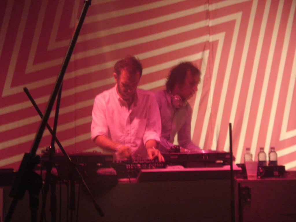 The two members of 2manydjs working a mixer and CDJs side by side under red stage light, in front of a striped backdrop