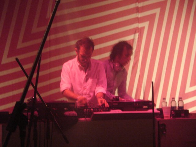 The two members of 2manydjs working a mixer and CDJs side by side under red stage light, in front of a striped backdrop