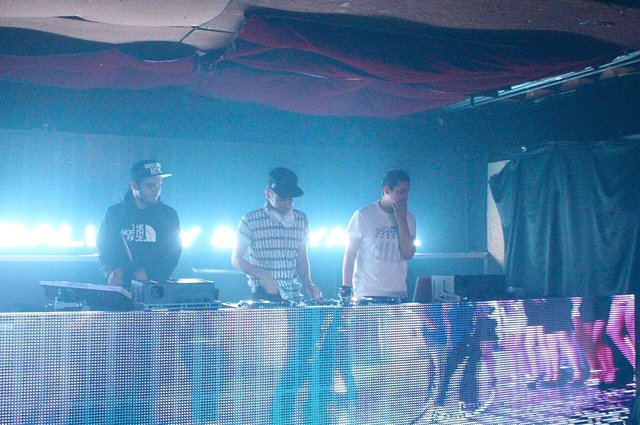 Three DJs standing behind a turntable booth lit by bright blue stage lights, with an LED panel in front
