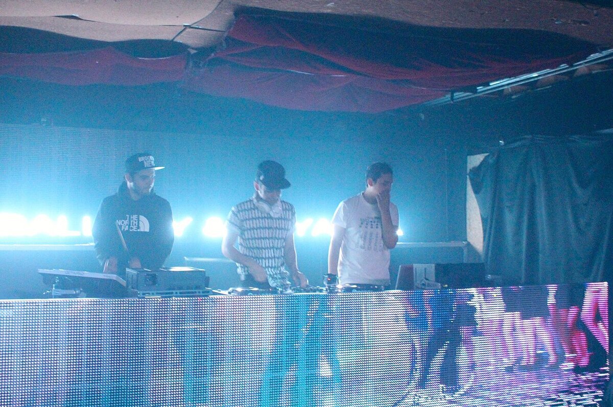 Three DJs standing behind a turntable booth lit by bright blue stage lights, with an LED panel in front