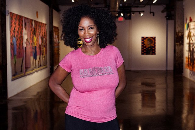 DJ Minx smiling in a pink t-shirt inside a gallery space with paintings on the walls