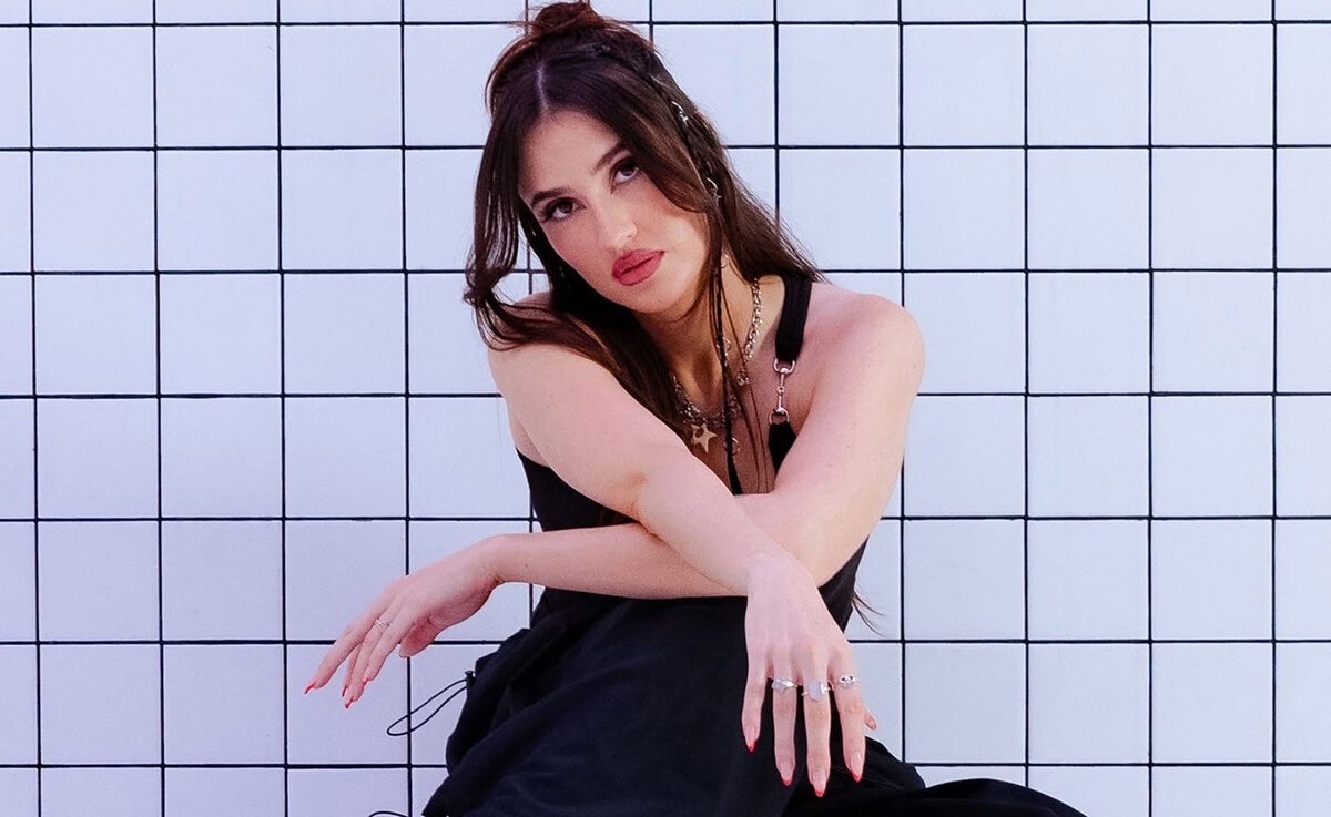 Portrait of a dark-haired artist in a black top posing against a white tiled wall