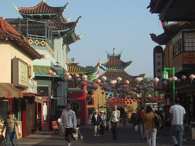 Pedestrians walking under rows of red and pink paper lanterns between pagoda-roofed buildings in Los Angeles Chinatown