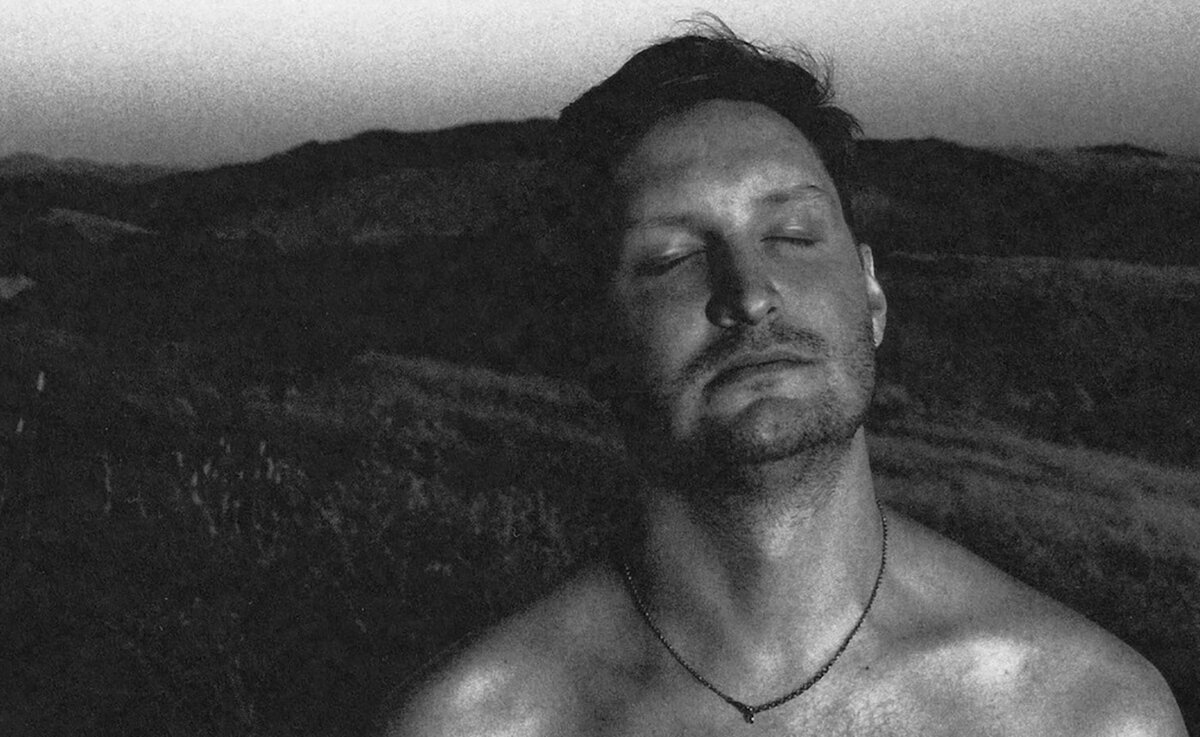 Black and white portrait of a man with his eyes closed and head tilted back against a dark hillside