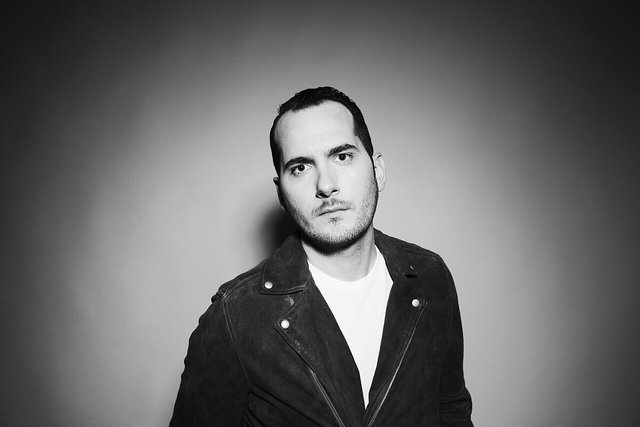 Black and white portrait of Andrew Bayer in a dark suede jacket and white t-shirt against a gray backdrop