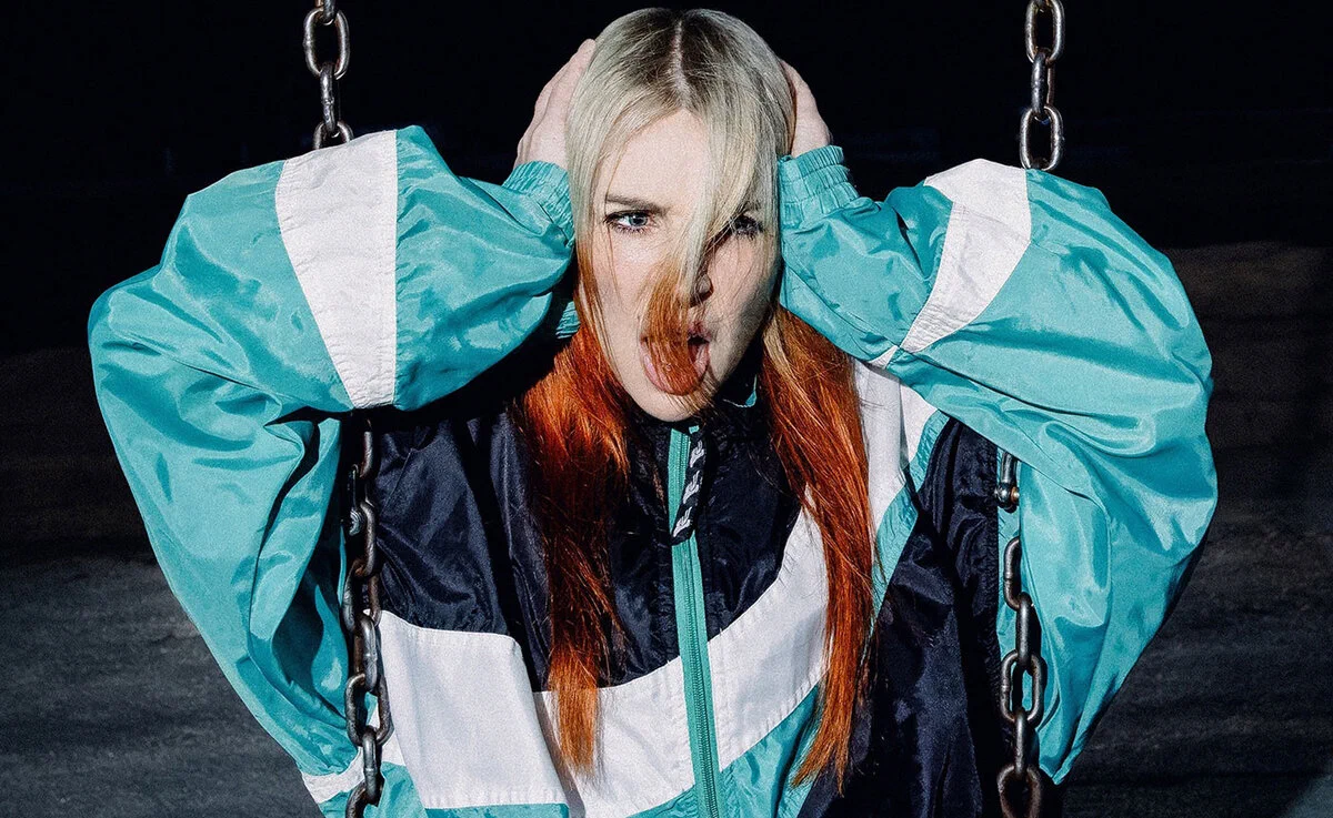 Alison Wonderland in a teal and orange jacket gripping swing chains in a promo portrait