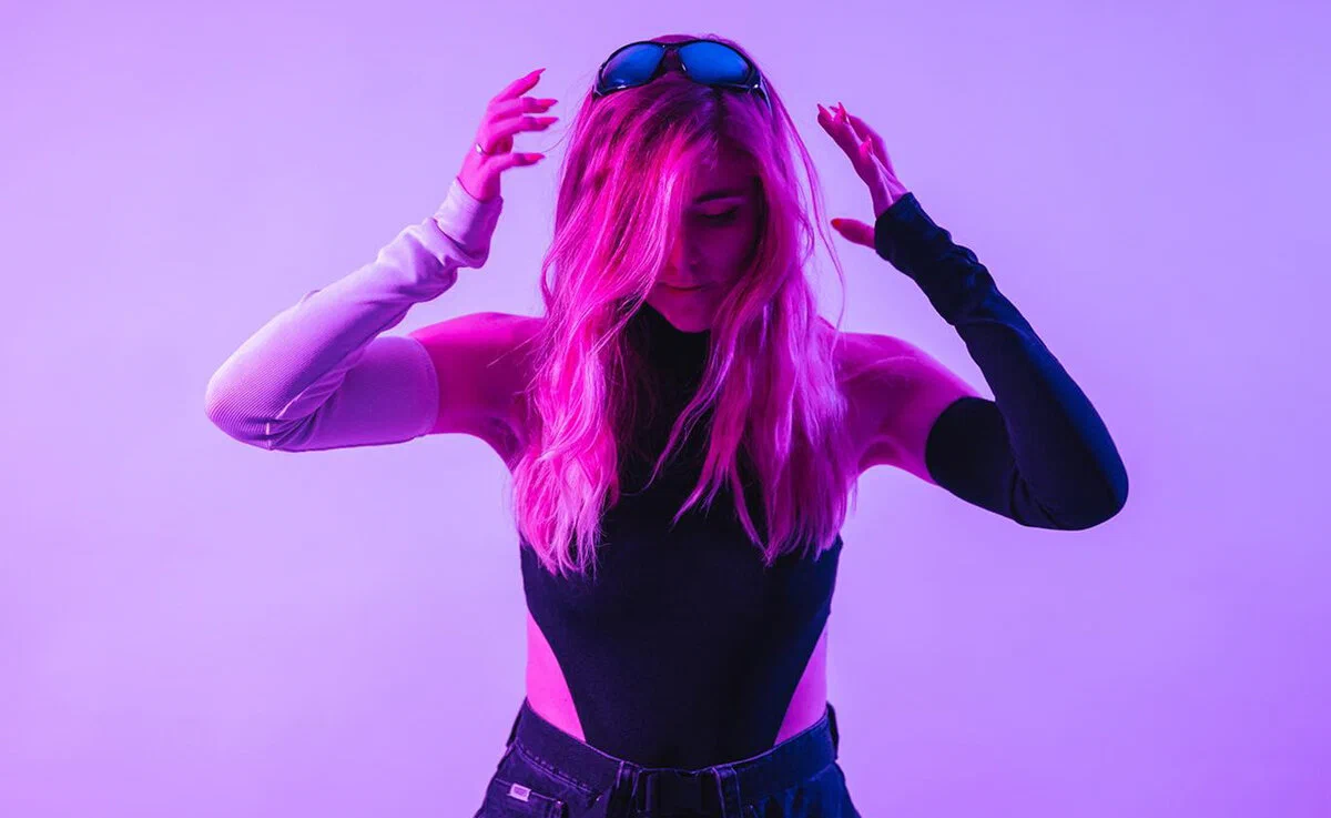 IVY with long pink hair against a purple backdrop, both hands raised near her head and sunglasses resting on top of her head