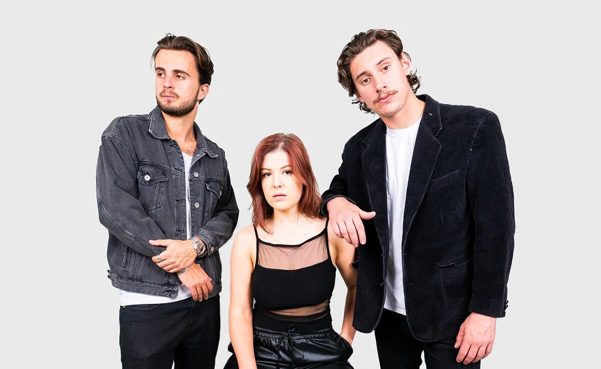 Three members of Laszewo standing against a plain light background, a man in a denim jacket and a man in a dark corduroy blazer on either side of a woman with red hair in a black top