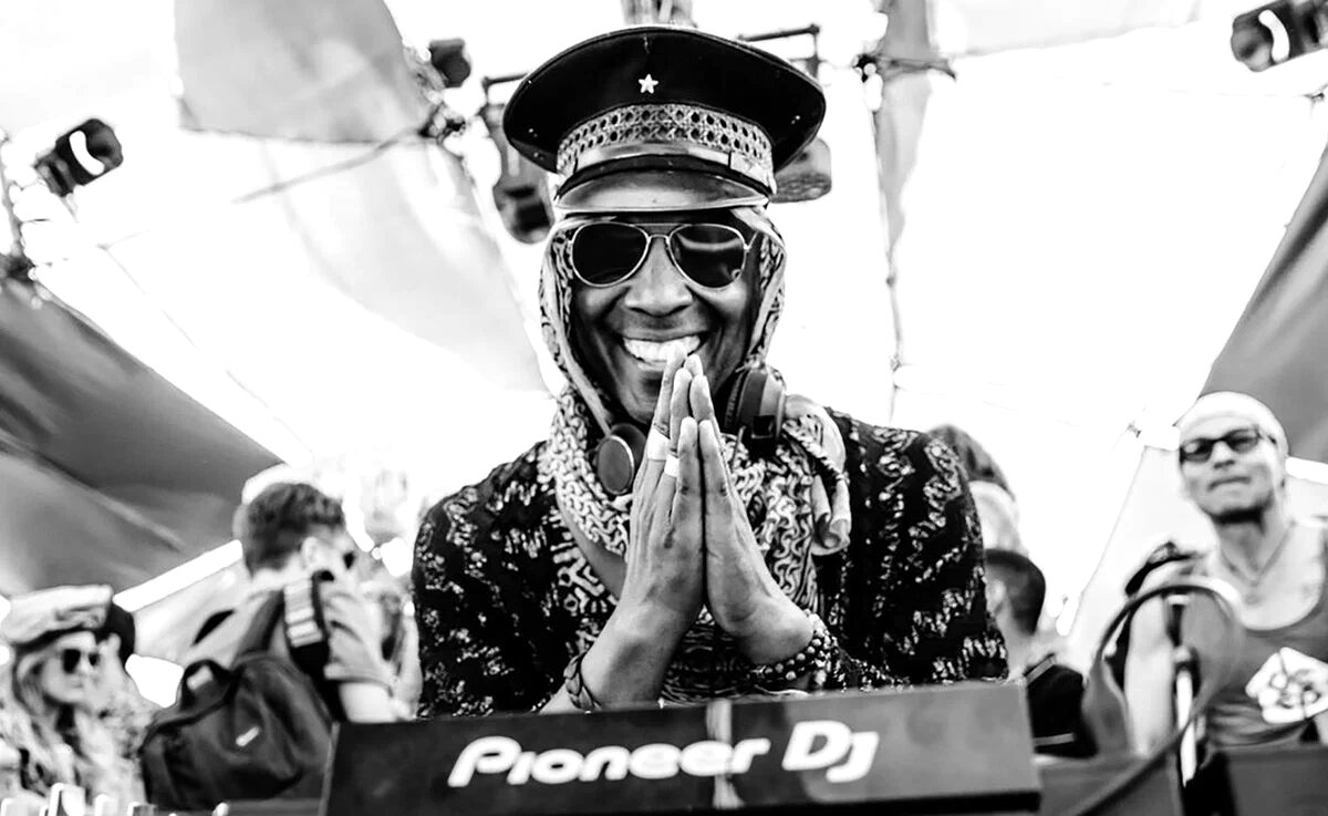 Black and white photo of Marques Wyatt smiling behind a Pioneer DJ deck with his hands pressed together, wearing a peaked cap, sunglasses and a patterned headscarf