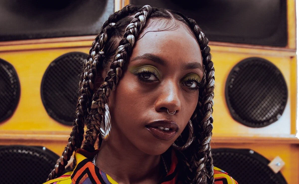 Nia Archives in a close-up portrait with braided hair and large hoop earrings, in front of stacked yellow speaker cabinets